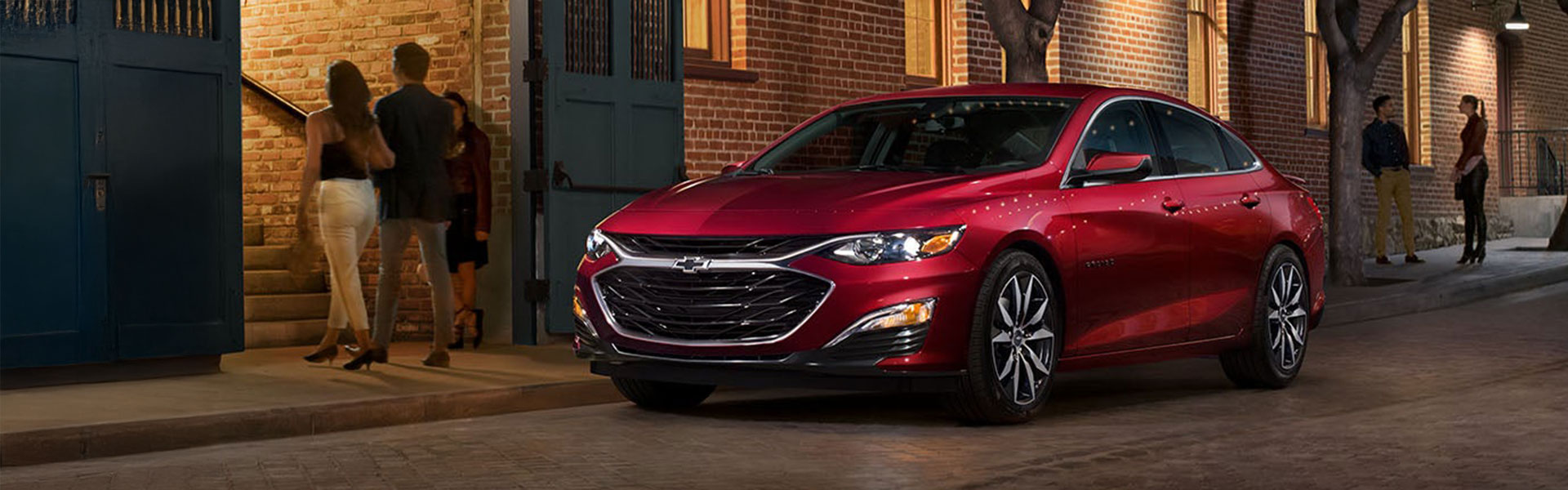 2025 Chevrolet Malibu red midsize sedan parked on a city street at night outside a brick building, highlighting its sleek exterior design