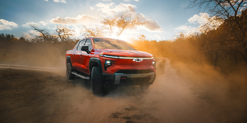 A red Chevrolet Silverado EV driving on a dirt road, kicking up dust with trees in the background.