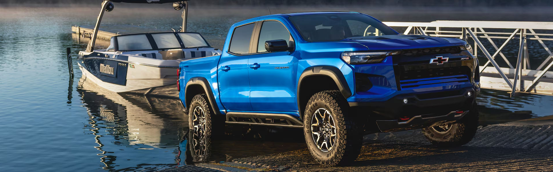 A blue 2026 Chevrolet Colorado parked near a motorboat on a calm lake at sunrise.